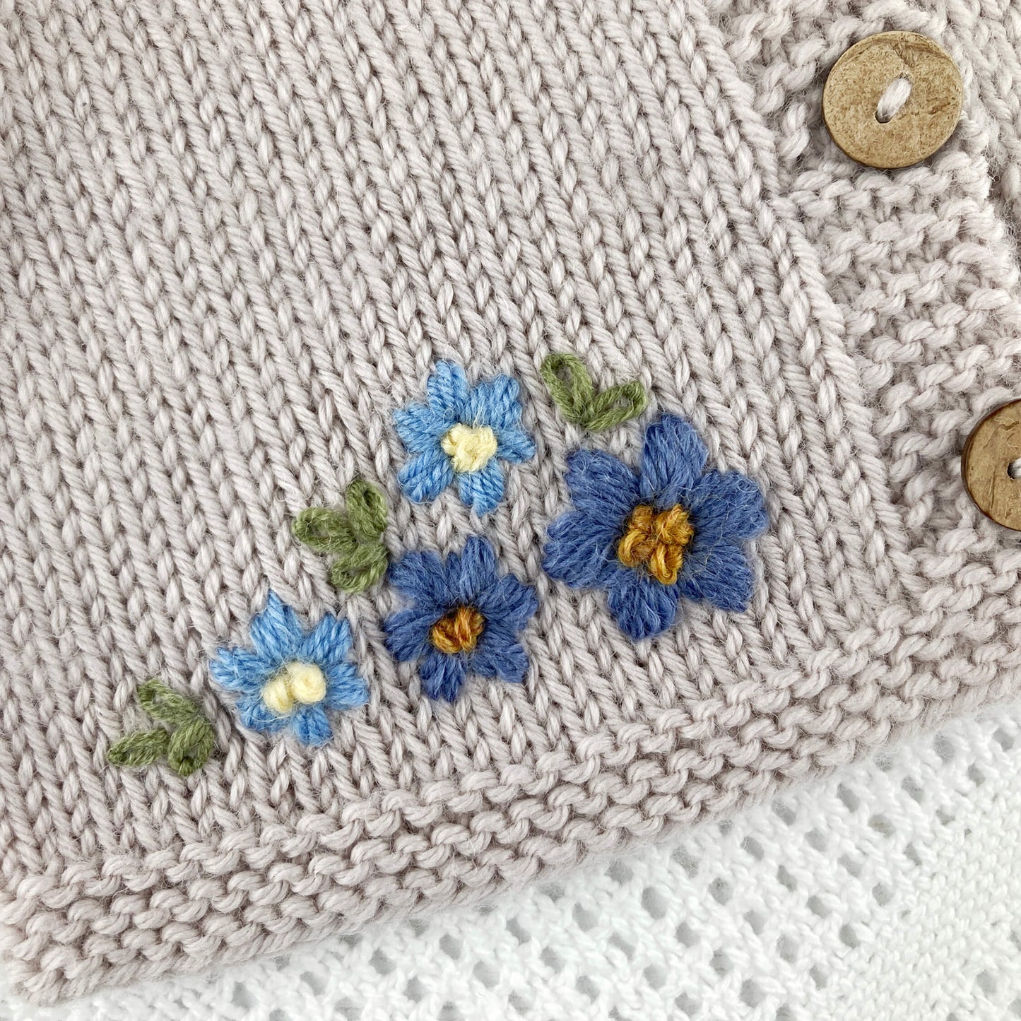 baby cardigan knitting pattern with embroidered blue flowers and buttons on a light grey background
