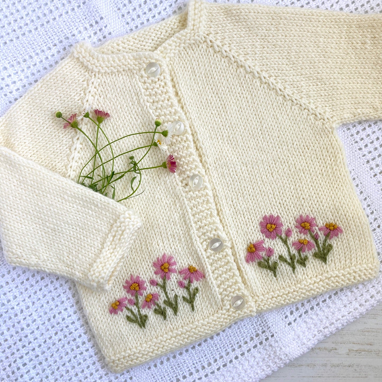 a hand embroidery pattern for baby clothing. daisy embroidery is featured along the bottom of a cream coloured baby cardigan