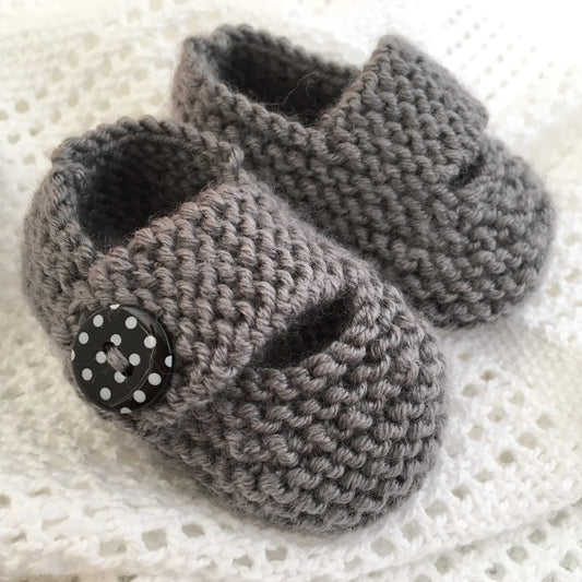 a baby shoes knitting pattern for beginners. Shoes have a wide strap and are knit in grey yarn with black spot buttons