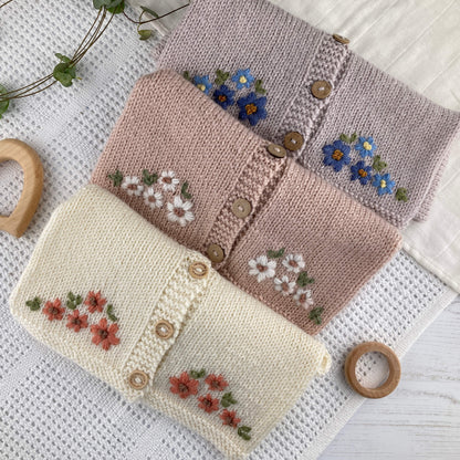 Knitting pattern for baby cardigan with floral pattern. 3 samples shown folded on a baby blanket