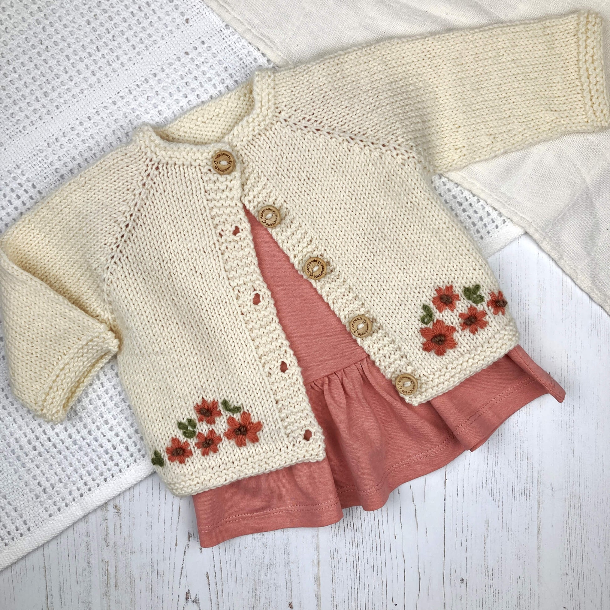 baby cardigan knitting pattern with floral embroidery shown over a pink dress on a white blanket