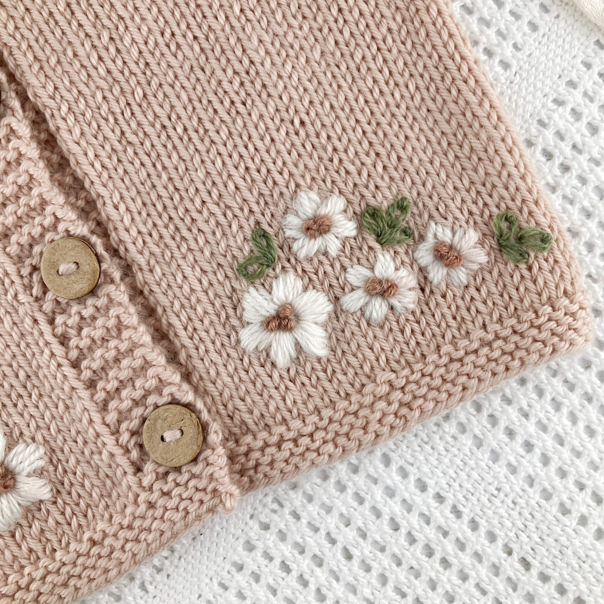 close up of baby cardigan knitting pattern with floral embroidery and buttons on a white blanket background