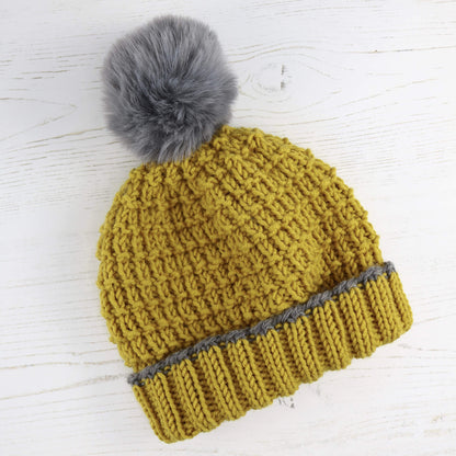 a hat knitting pattern in family sizes. sample shown is knit in mustard colour yarn with a faux fur pompom