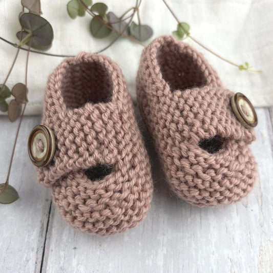 pdf knitting pattern for baby booties. Sample is shown knit in garter stitch in dusky pink yarn the brown and beige buttons sitting on whitewashed wooden floor with greenery in the background