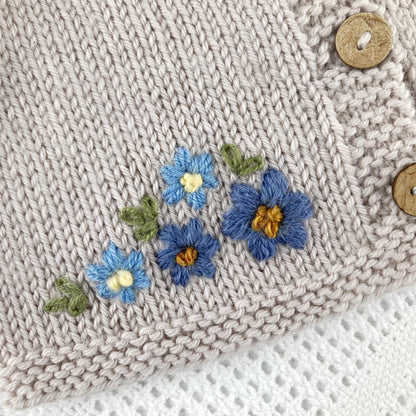 baby cardigan knitting pattern with embroidered blue flowers and buttons on a light grey background